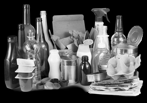 A collection of various discarded household items and packaging materials arranged on a flat surface, including glass bottles of different shapes and sizes, plastic bottles and containers, metal cans, and a stack of newspapers or paper documents. Some items are partially obscured by others, with a cardboard box visible in the background holding more debris. The scene appears to be an indoor or outdoor setting for rubbish collection, highlighting typical waste materials that a commercial waste removal service like Commercial Waste Pinner might handle in Pinner, Middlesex. The objects vary in texture, from smooth glass and plastic to crinkled paper and metallic surfaces, with the overall composition suggesting an accumulation of recyclable waste ready for disposal or collection. The lighting is even, clearly illuminating all objects on the surface, providing a detailed view suitable for accessibility and SEO purposes related to rubbish removal services in the local area.