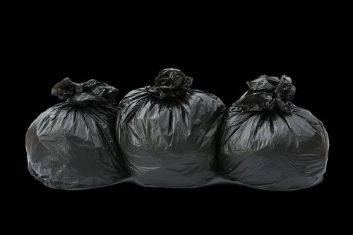 Three large black rubbish bags made of plastic and tightly tied at the tops are arranged in a row on a dark, featureless background. The bags appear to be filled with waste materials, with visible creases, folds, and slight bulges indicating bulky contents. The surface texture of the bags is shiny, reflecting minimal light, which highlights their crinkled appearance. The bags are positioned on what seems to be an outdoor or industrial surface, possibly a driveway or pavement, although the background provides no environmental context. This image is relevant to rubbish removal services in Pinner, showing typical waste collection packaging awaiting collection by a professional waste management company, such as Commercial Waste Pinner. The scene emphasizes the act of waste disposal, with focus on the black plastic bags used for household or commercial waste storage, suitable for local SEO targeting customers needing rubbish clearance in the area.