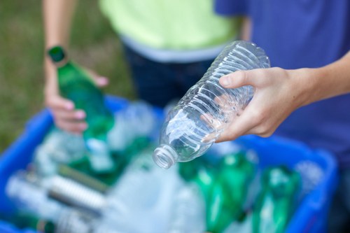 A close-up view of a person's hand holding a clear plastic bottle in the foreground, with a blue recycling bin filled with crushed plastic bottles and containers in the background. The person's arm is visible, with light skin and wearing a short-sleeved shirt. Behind them, another individual wearing a green high-visibility vest is seen, holding a green plastic bottle. The scene appears to take place outdoors on a grassy area, possibly as part of a waste collection or recycling activity in Pinner, with natural daylight illuminating the scene. The focus is on the plastic bottles and recycling effort, consistent with rubbish removal services offered by Commercial Waste Pinner.