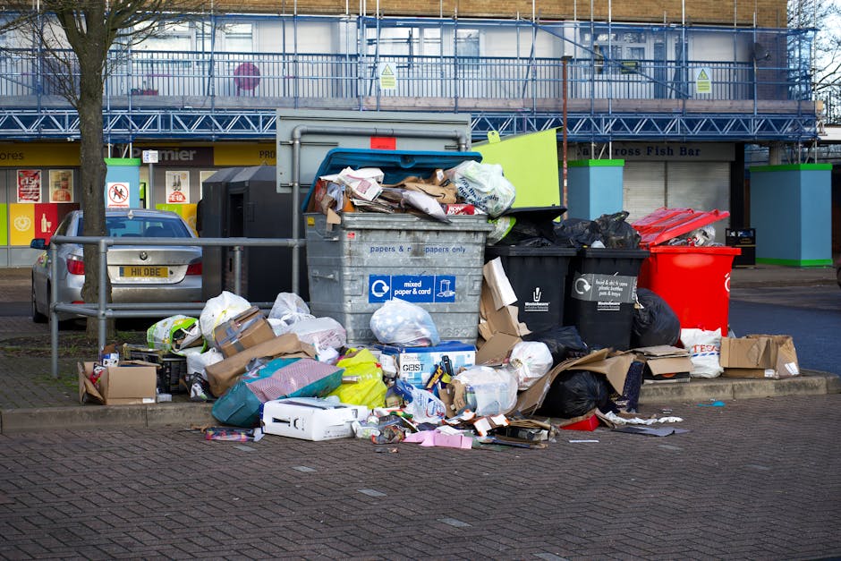 A pair of black plastic rubbish bags, filled and tied at the top, are placed on the edge of a paved sidewalk in an outdoor urban setting. The bags appear to contain household waste and are positioned 