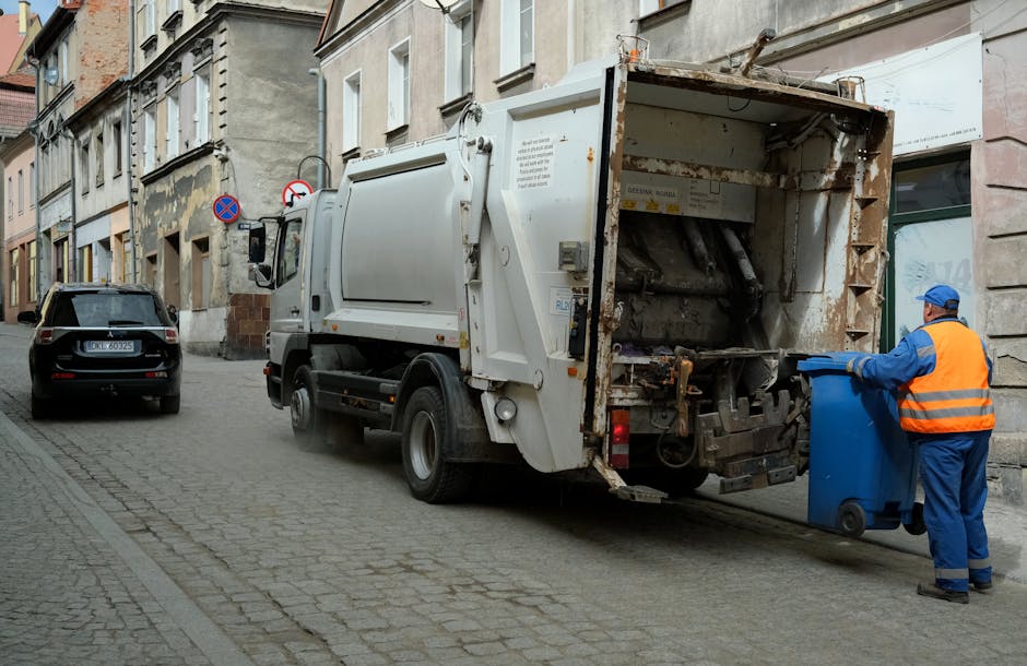 A person wearing a dark hoodie, gray beanie, and high-visibility vest stands on a modern train station platform beside a red-framed, hand-pulled trolley filled with various types of waste materials, i